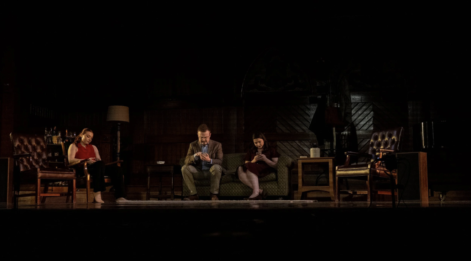 Three people sit on a sofa and chairs in a dimly lit room, all looking at their phones. The room has dark wood paneling and some furniture, including a lamp, a table, and a bar area with bottles.