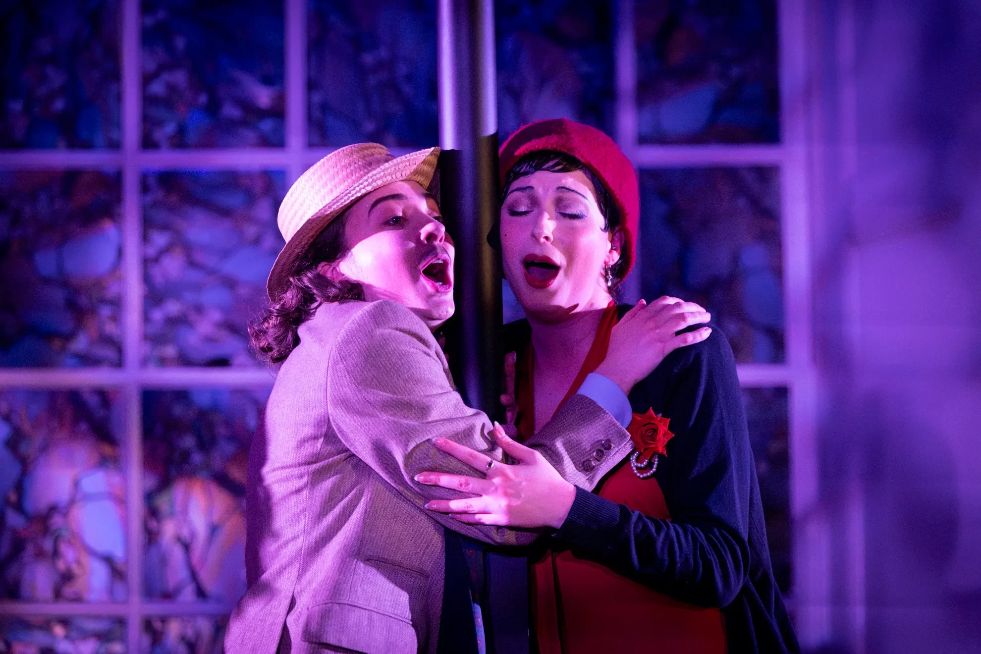 Two women passionately singing together, one dressed in a beige blazer and fedora, the other in a red hat and dress, in a dimly lit room with purple lighting and a stone wall background.