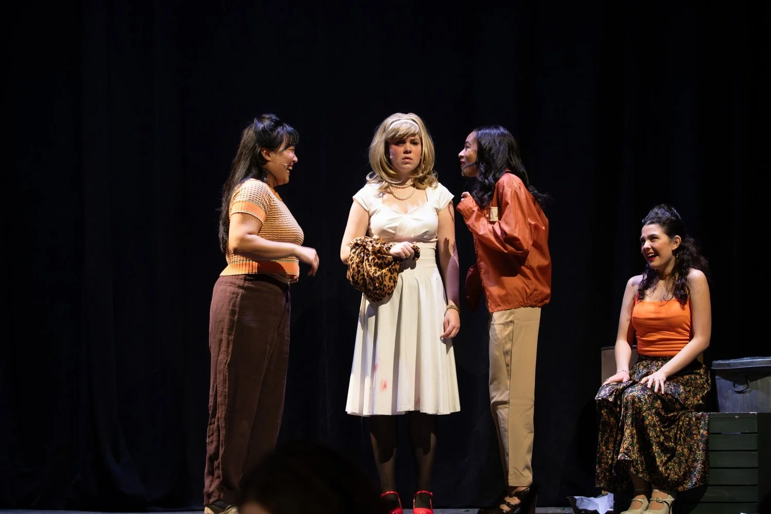 Five women on stage in a theatrical performance with black background; one woman is sitting while the other four are standing, engaging in conversation.