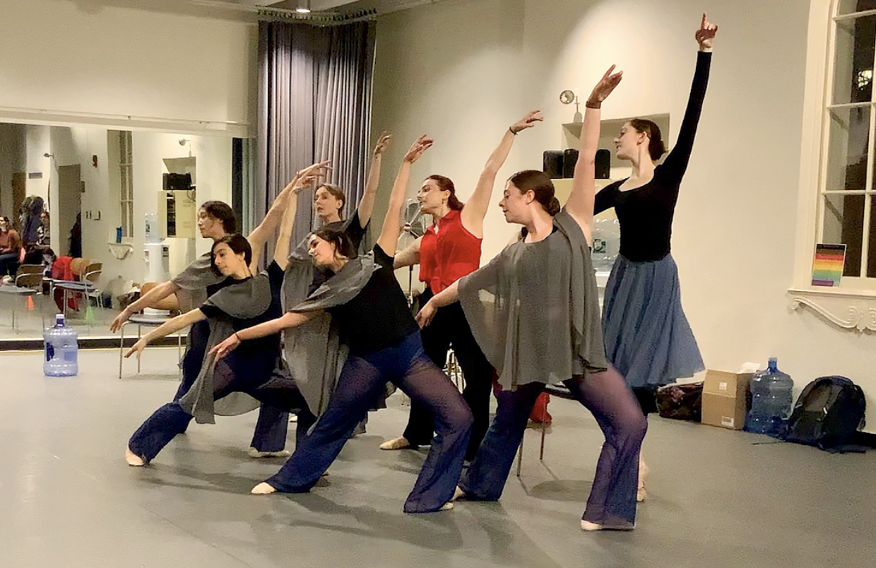 Group of seven women dancing in a studio, with some women in front and others in back, all with arms raised in graceful poses, wearing casual dance attire.