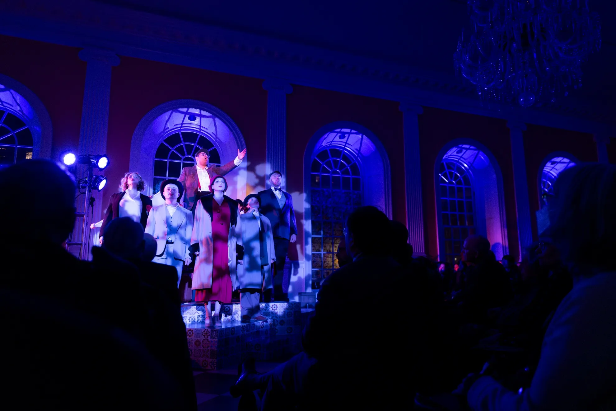 People performing on stage in a brightly lit, elegant room with large arched windows and chandeliers.