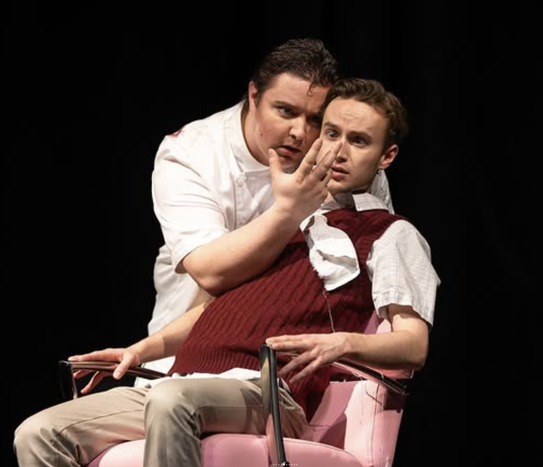 Two actors on stage, one dressed as a nurse and the other as a patient, engaging in a dramatic scene.