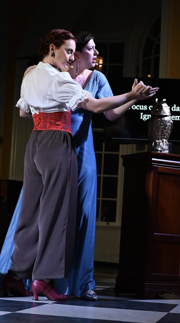 Two women in vintage style clothing standing closely at a dark stage, indicating a theatrical performance or presentation.