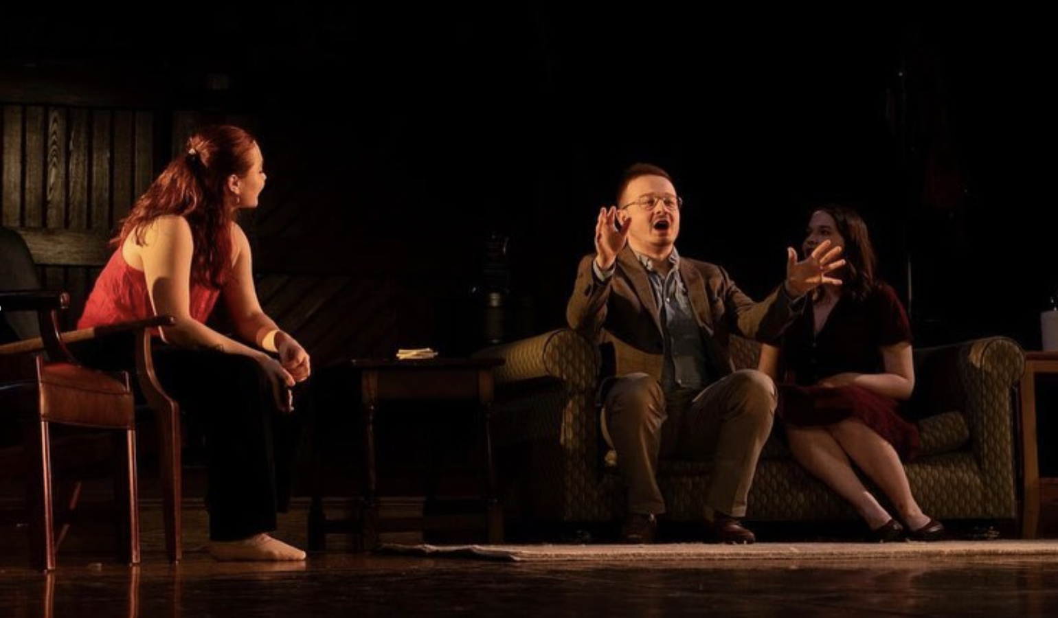 Three actors performing in a theatrical play on stage, with one woman sitting on a chair and two on a sofa, engaged in a scene with expressive gestures.