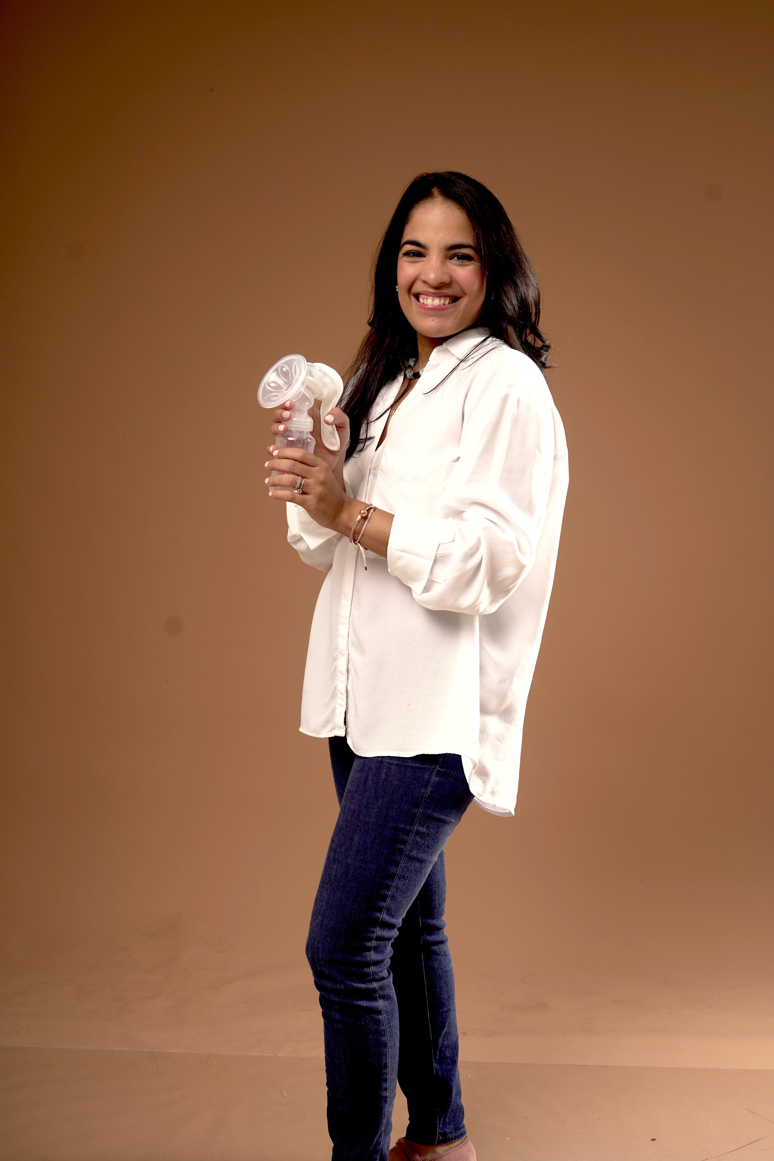 A woman with dark hair smiling while holding a white handheld fan, wearing a white shirt and dark jeans, standing against a plain brown background.