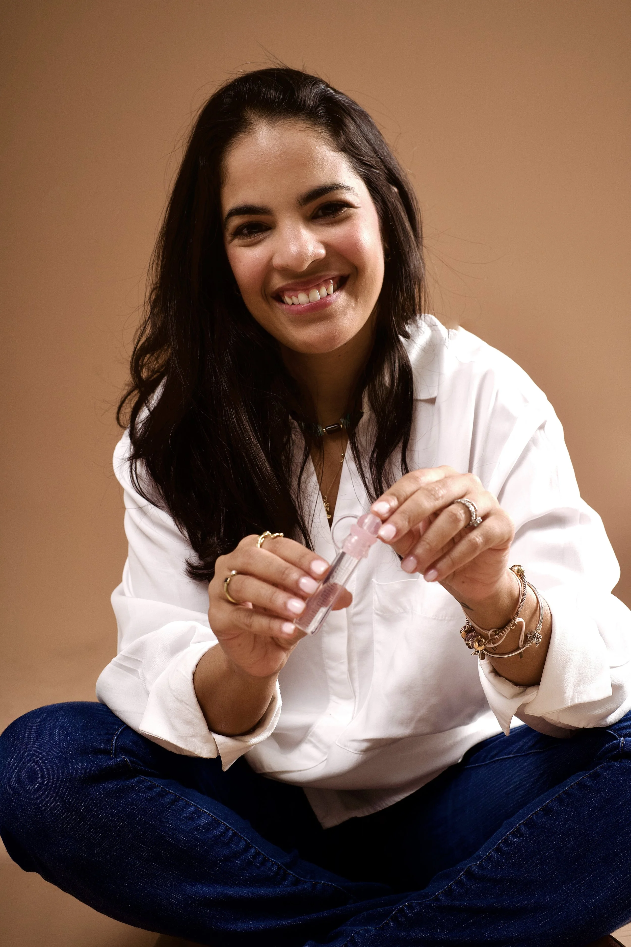 Woman with long dark hair seated cross-legged, holding a small vial with a syringe, smiling at the camera, wearing a white blouse and blue jeans, in front of a plain beige background.