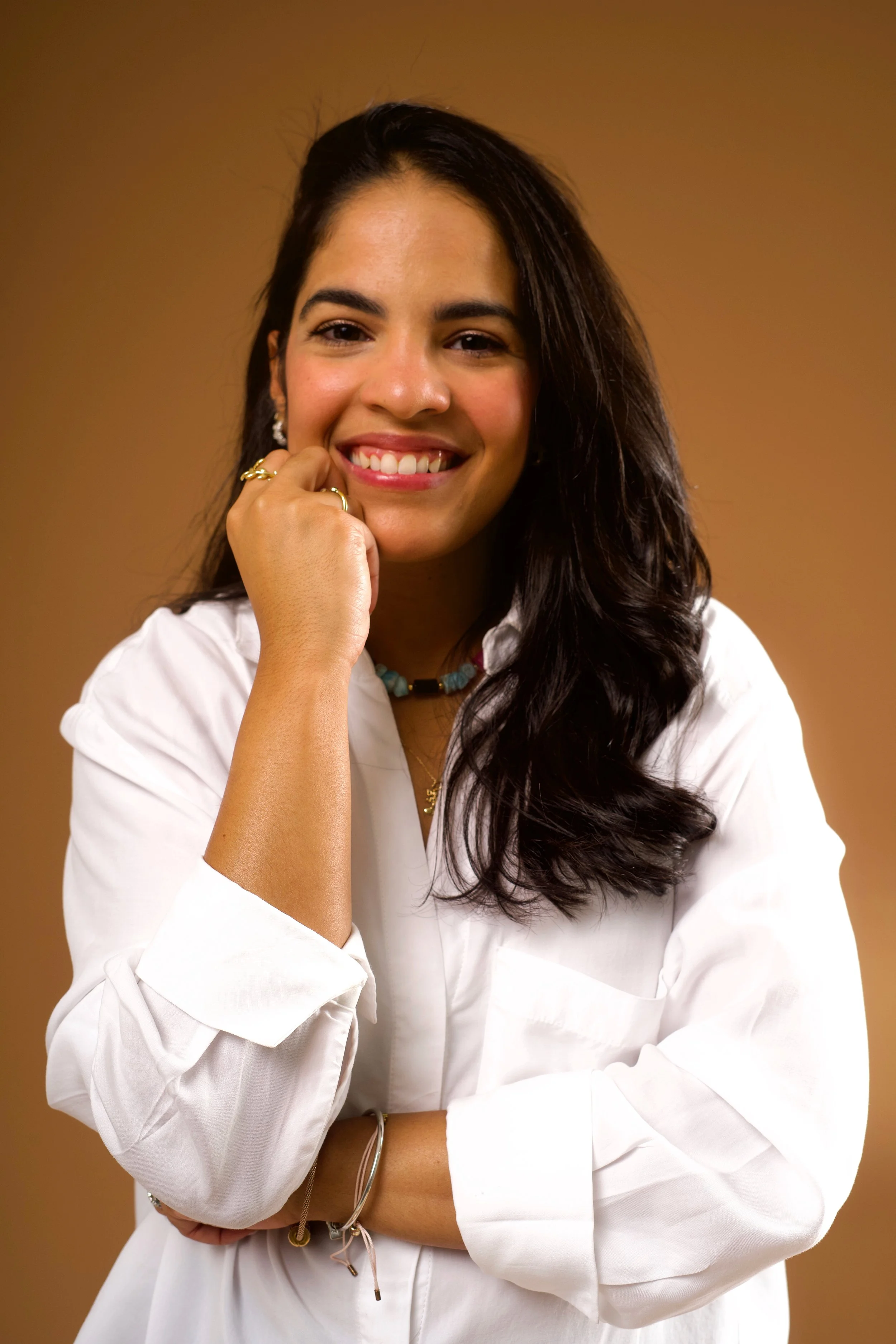 Mujer joven sonriendo, con cabello oscuro y largo, usando ropa blanca y accesorios de joyería, con fondo marrón.