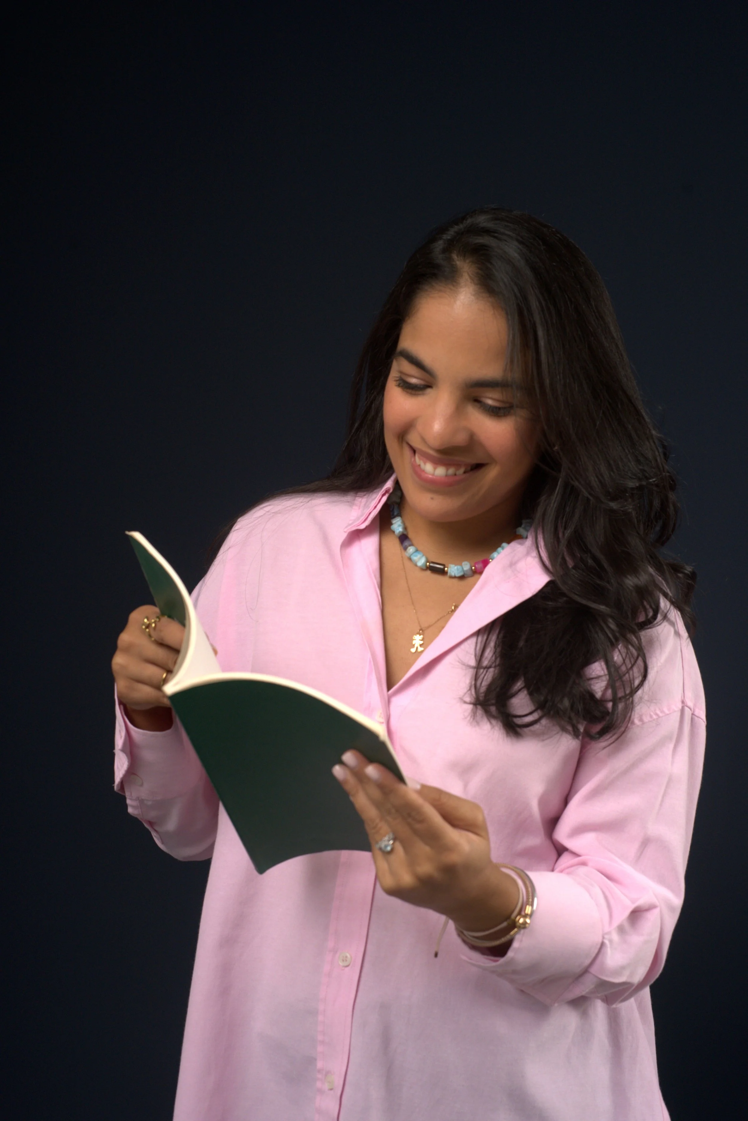 A woman with long dark hair wearing a pink shirt and multicolored beaded necklace, smiling while reading a book against a dark background.