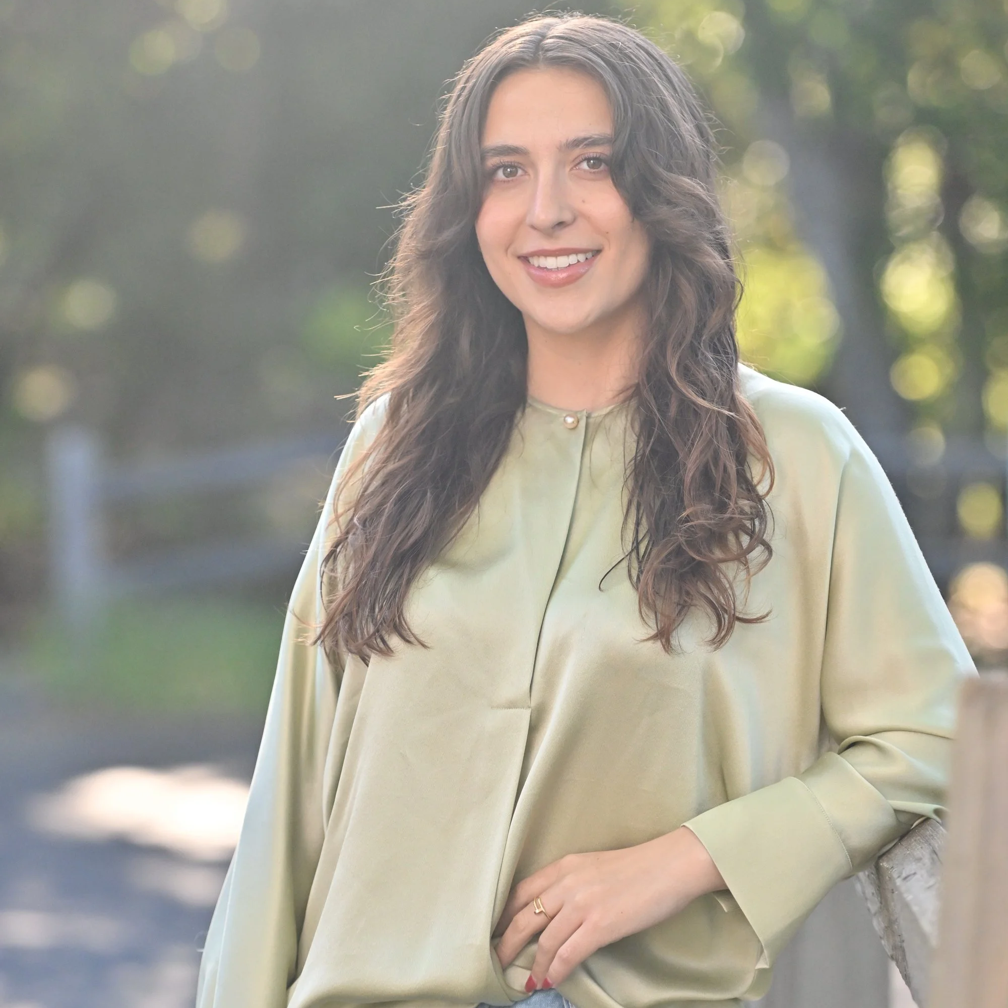 A young woman with long, wavy brown hair, smiling with teeth visible, wearing a light green shirt with a collar and pearl detail, outdoors with a blurred background of trees and sunlight.