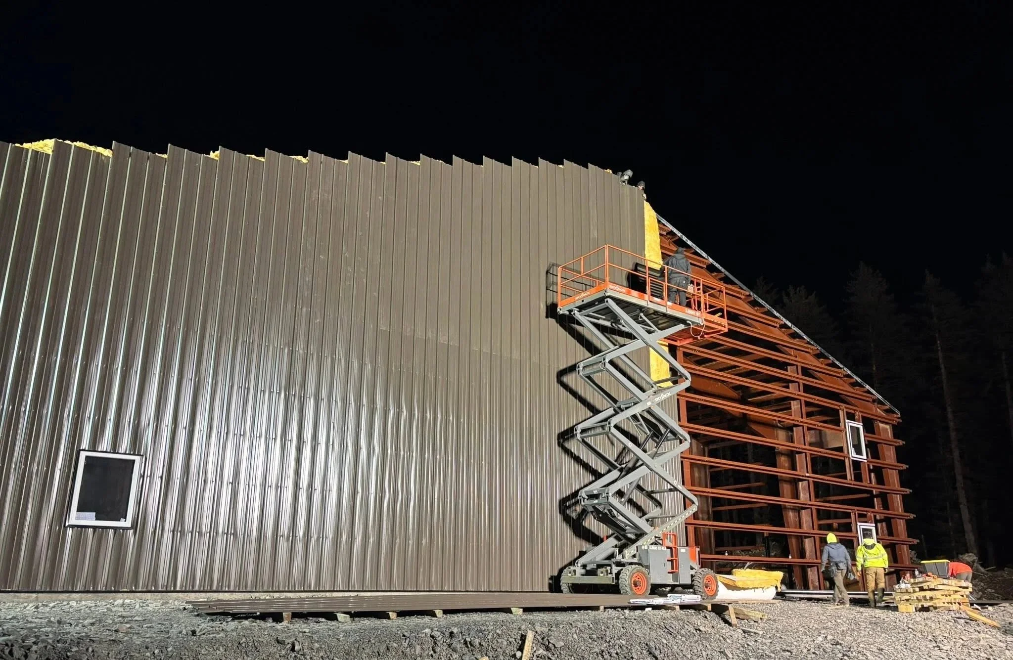 Construction crew installing metal siding on a large steel-frame building at night using a scissor lift.