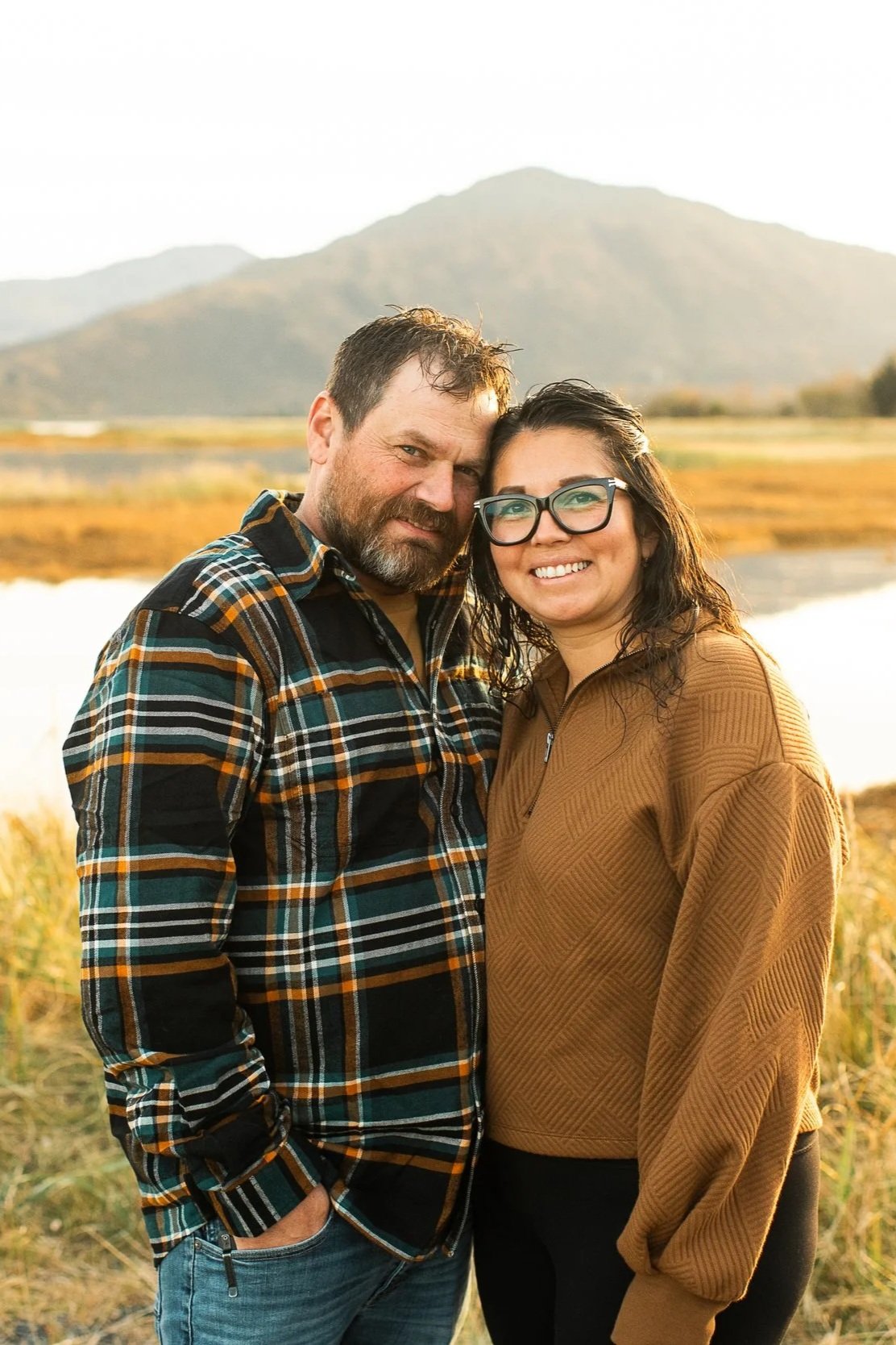 A man and a woman standing outdoors near a body of water with mountains in the background, smiling at the camera.