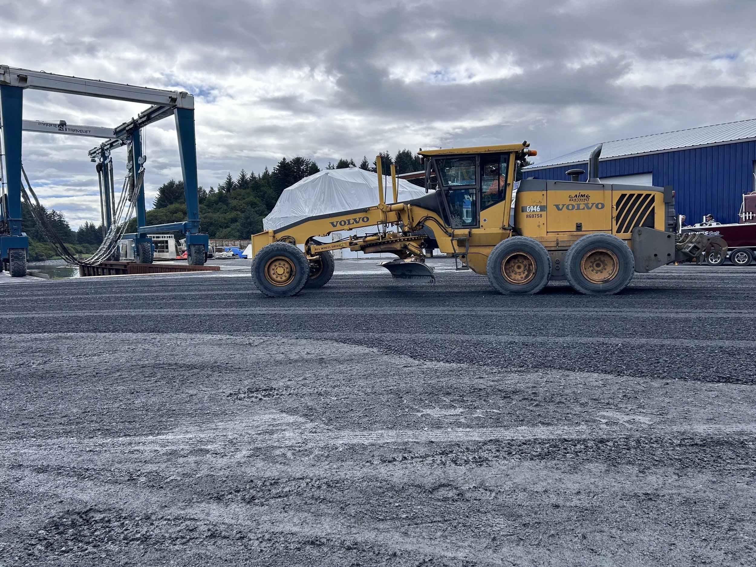 A yellow Volvo construction vehicle with black stripes on the side and a large scoop at the front, parked on a textured black surface. In the background, there is a blue building, some cars, and trees along a hilly landscape. The sky is cloudy.