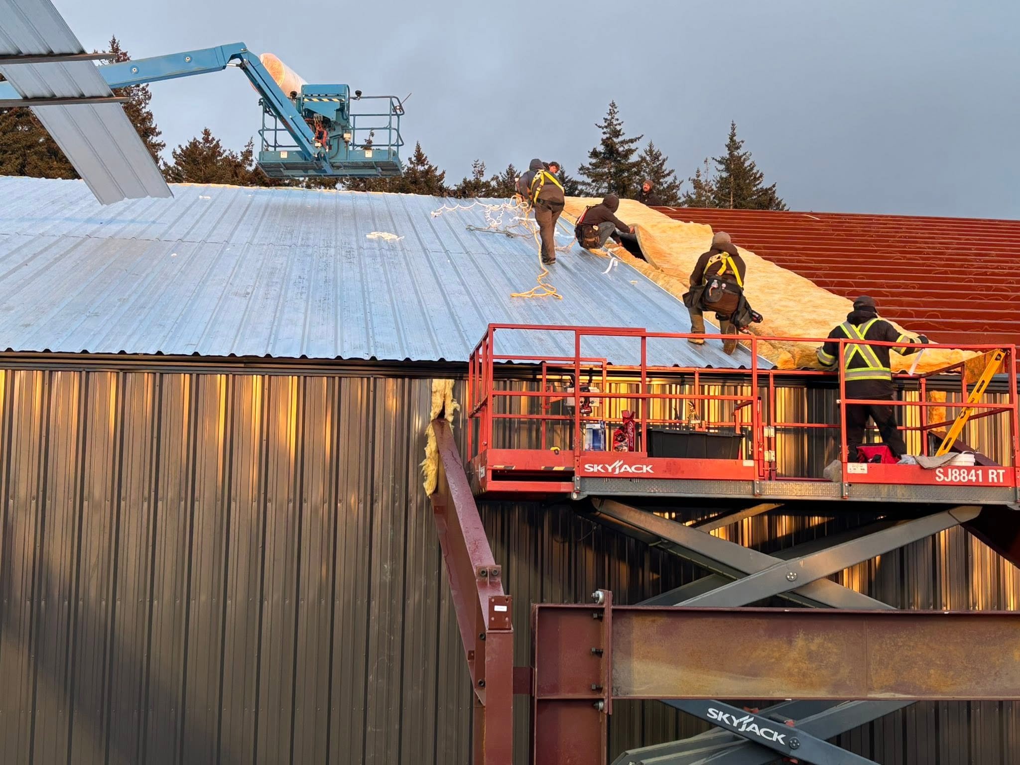 Construction crew installing insulation and metal roofing on a large steel building using scissor and boom lifts.