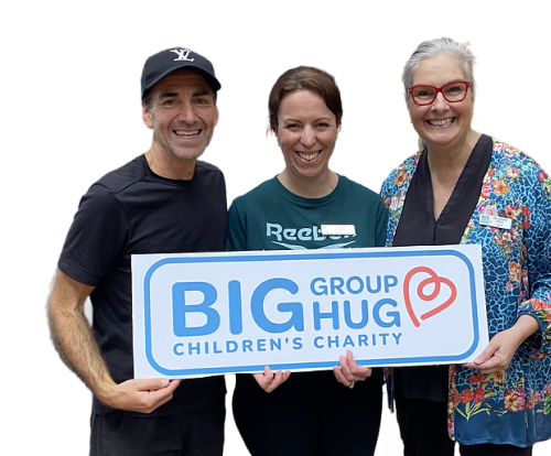 Three people standing together, smiling, holding a large sign that reads "BIG GROUP HUG CHILDREN'S CHARITY." The person on the left is a man wearing a black shirt and a black baseball cap. The middle person is a woman with short brown hair wearing a green shirt. The person on the right is a woman with gray hair, glasses, wearing a colorful patterned jacket.