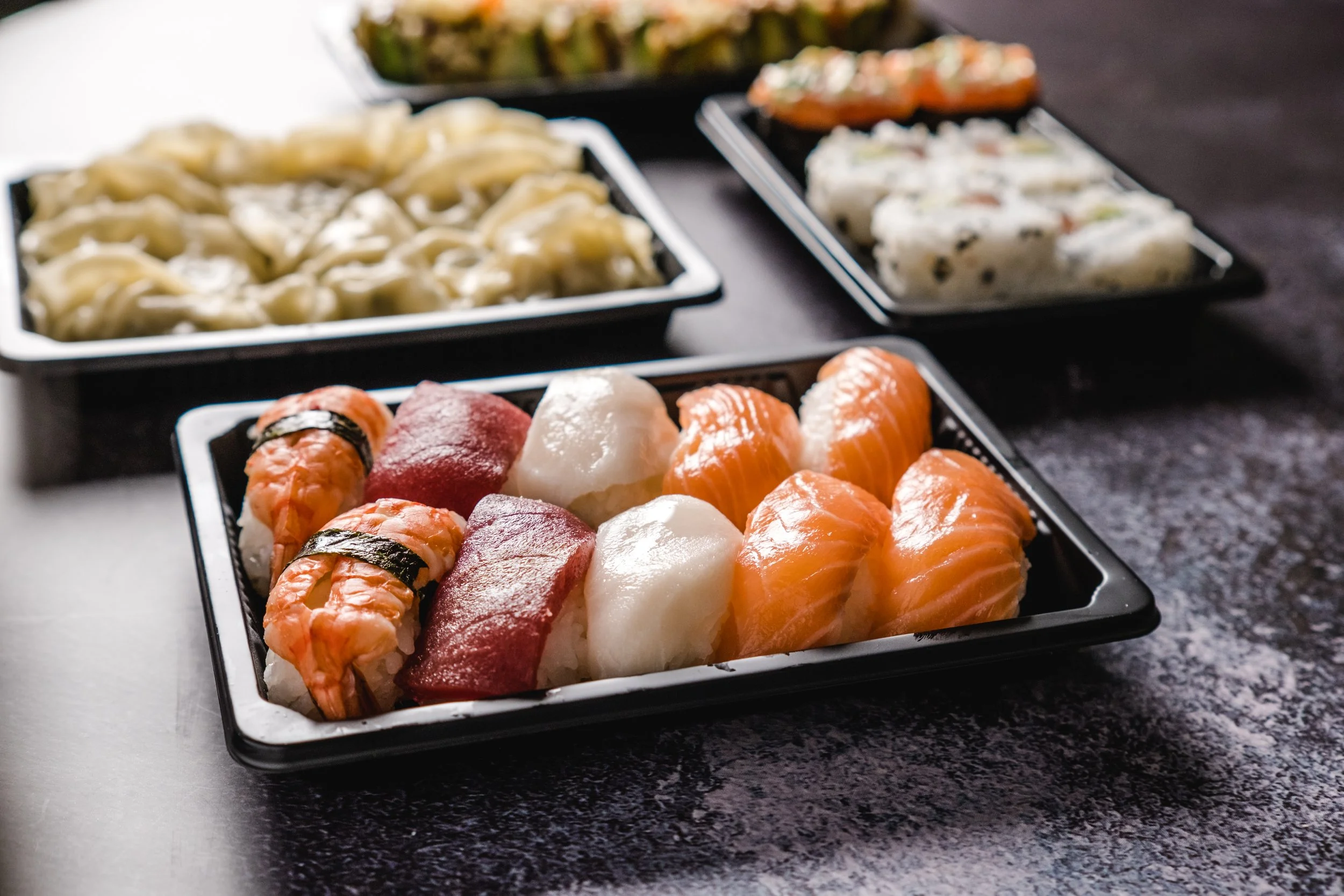 Assorted sushi rolls and sashimi on black trays, including salmon, tuna, and other fish, with rice and seaweed, arranged on a dark textured surface.