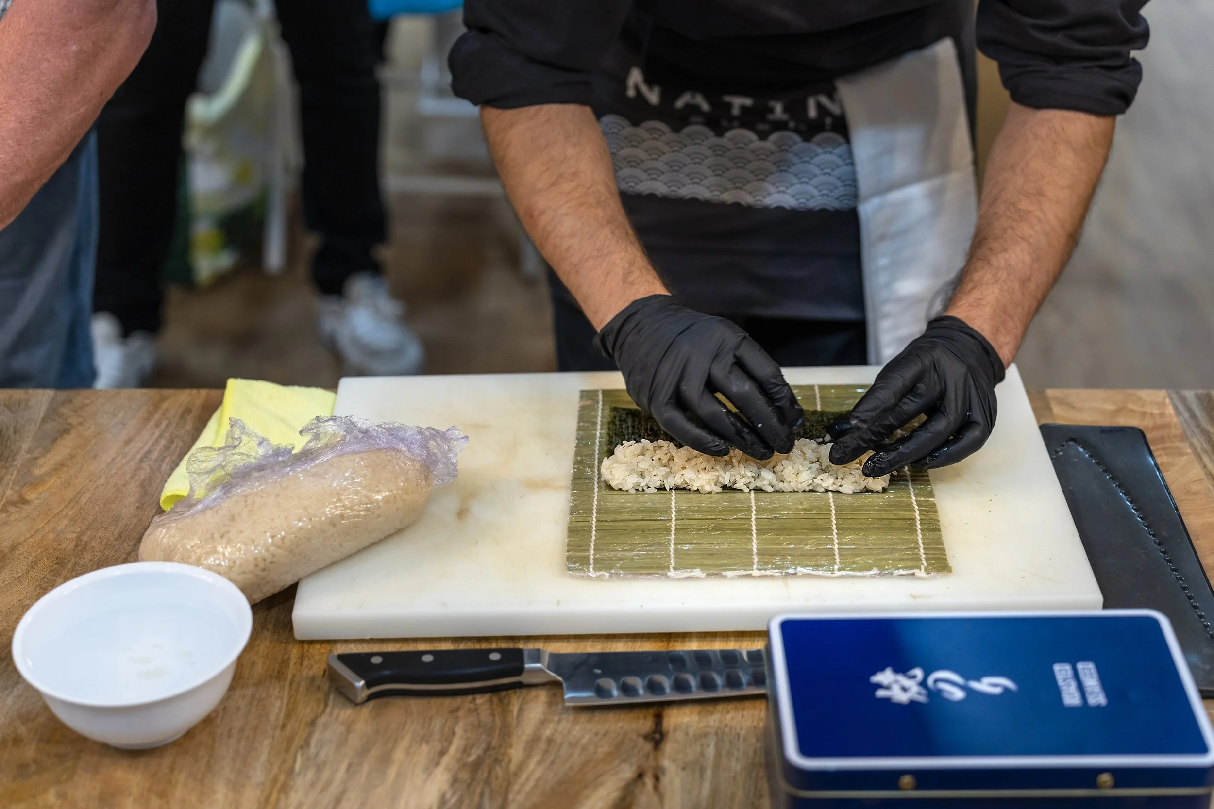 A person wearing black gloves is preparing sushi on a bamboo mat, with ingredients including rice and seaweed. A packaged piece of raw fish, a white bowl, and a knife are on the wooden table.