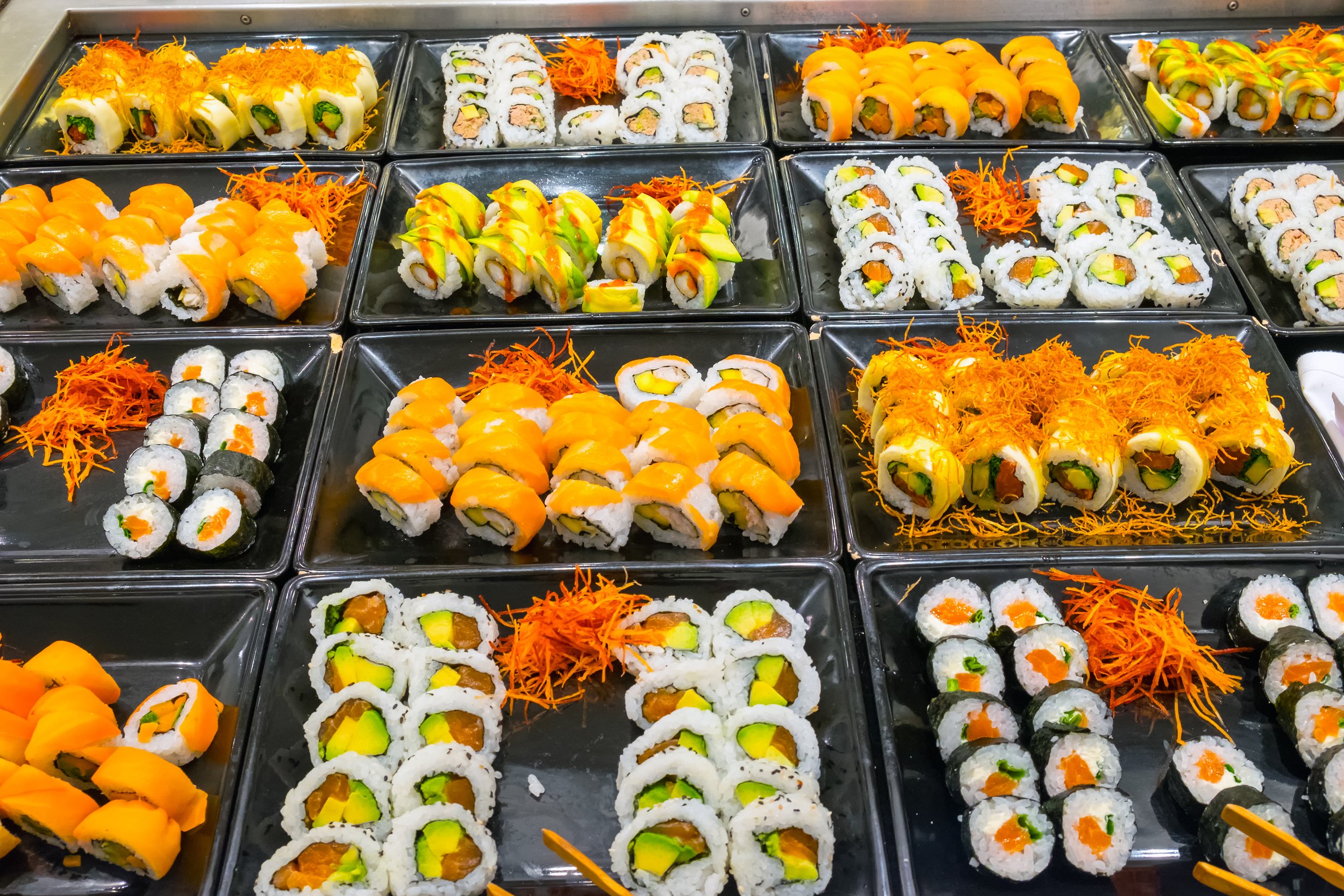 Assorted sushi rolls on black trays, including rolls with salmon, avocado, cucumber, and crab meat, garnished with shredded carrots and drizzled sauces, arranged in rows on a stainless steel counter.