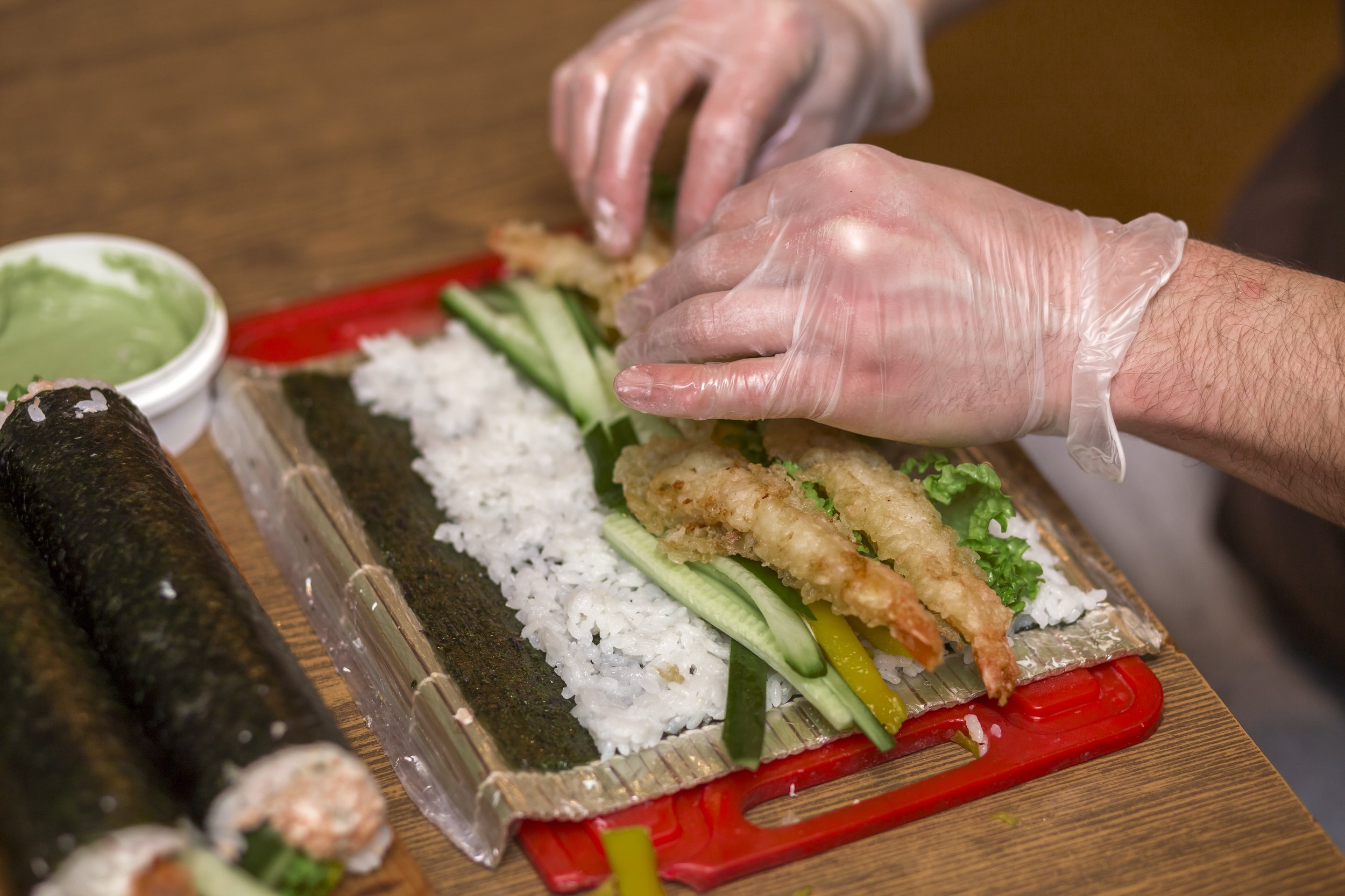 Hands wearing clear gloves preparing to make sushi on a bamboo mat. Sushi ingredients can be seen, including sushi rice, cucumber, red bell pepper, and fried fish fillet. Rice, seaweed wrap, and sushi rolls are visible on the table.