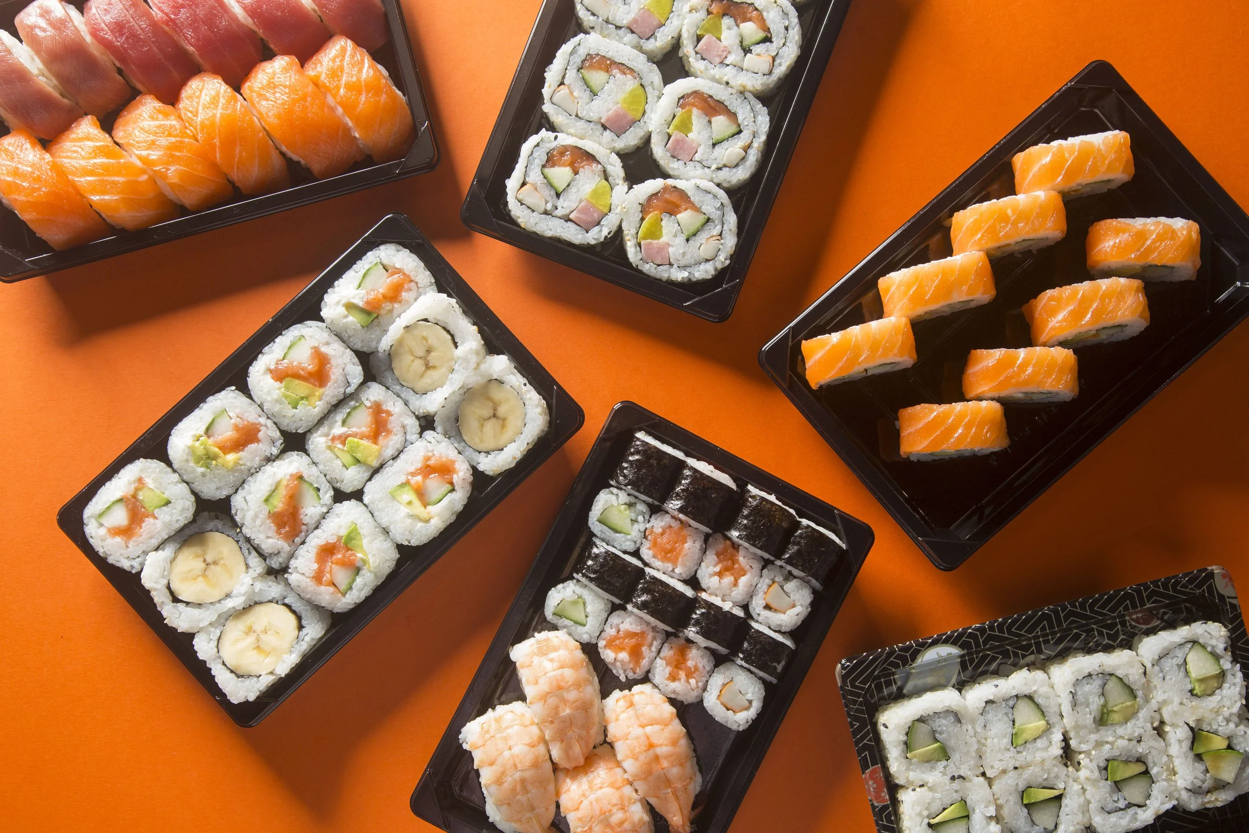 Assorted sushi rolls on black trays placed on an orange surface, including salmon nigiri, sushi with banana, and various types of maki rolls with vegetables and fish.