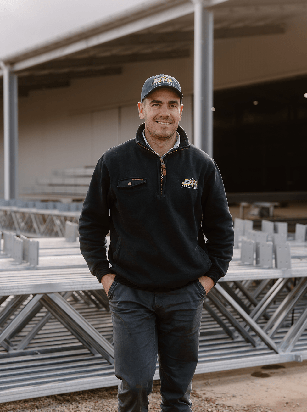 A man in casual work attire, including a hat and jacket with the logo 'BIG Steel'. He is standing outdoors in front of metal bleachers or scaffolding, smiling with his hands in his pockets.