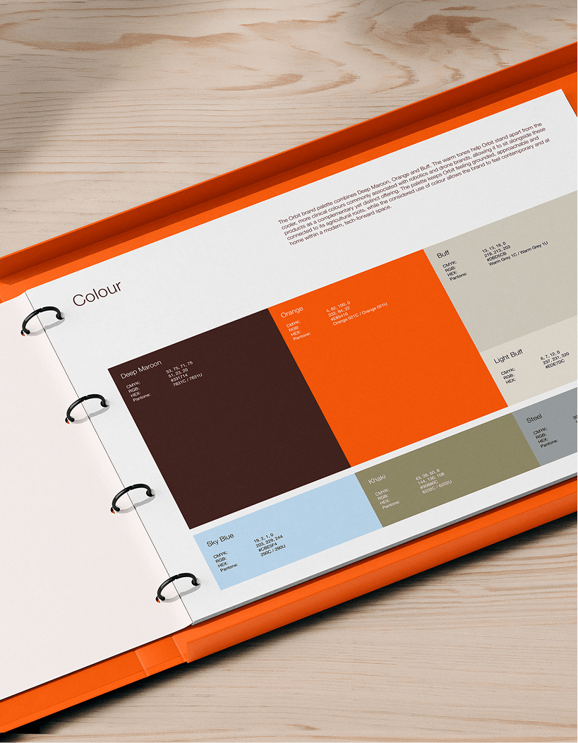 A color sample book open on a light wood surface, displaying various paint color swatches including Deep Maroon, Orange, Sky Blue, Khaki, and Steel, with descriptions and codes.