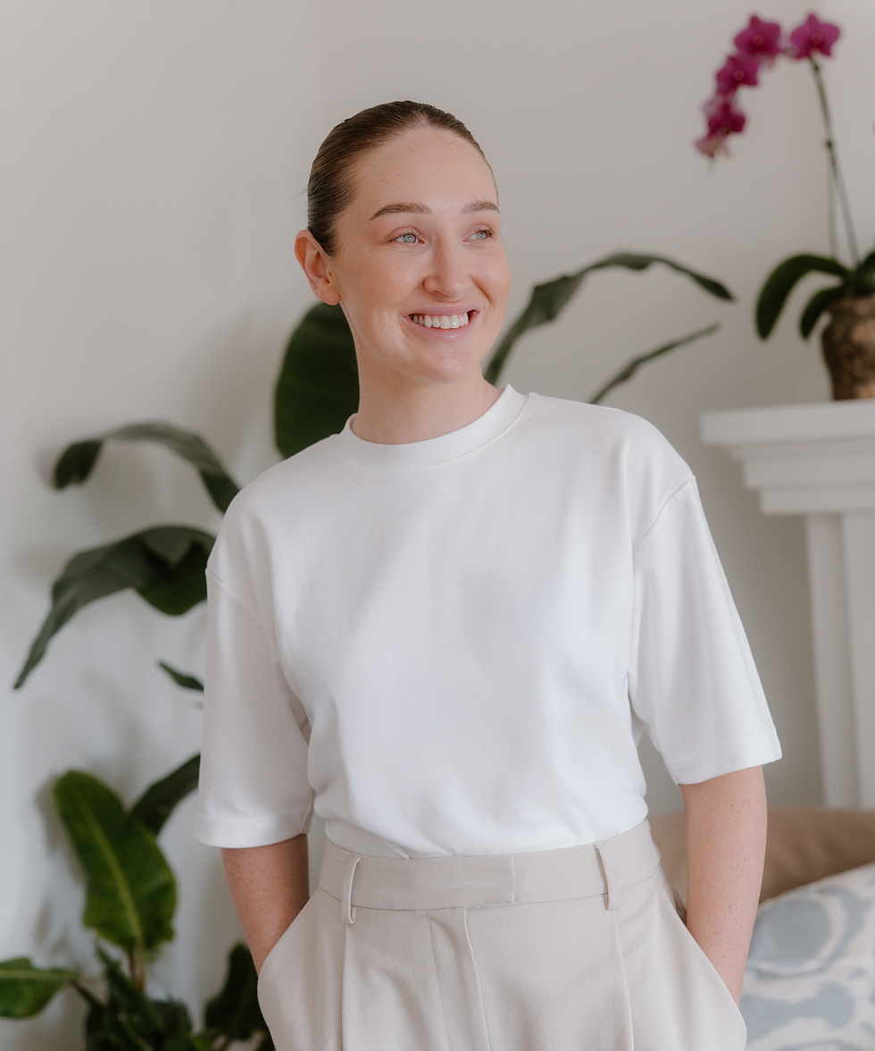 A young woman with fair skin and brown hair tied back, smiling and looking to her left, wearing a white t-shirt and beige pants, standing in a room with green plants and a white fireplace mantel with a potted orchid on it.