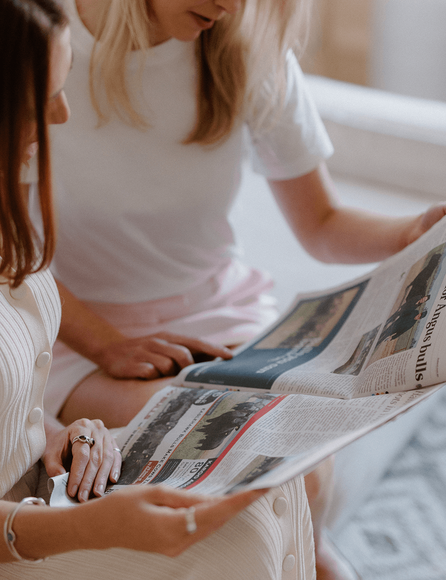 Two women sitting on a bed reading a newspaper together.