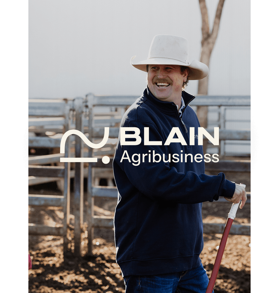 A man wearing a wide-brimmed white hat and a navy blue jacket standing outdoors on a farm, smiling, holding a shovel, with wood fencing and a tree in the background.