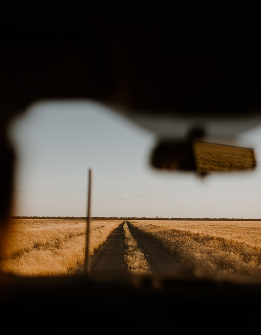 View through a dusty car windshield of a rural dirt road stretching into the distance, flanked by fields of tall golden grass, under a clear sky.