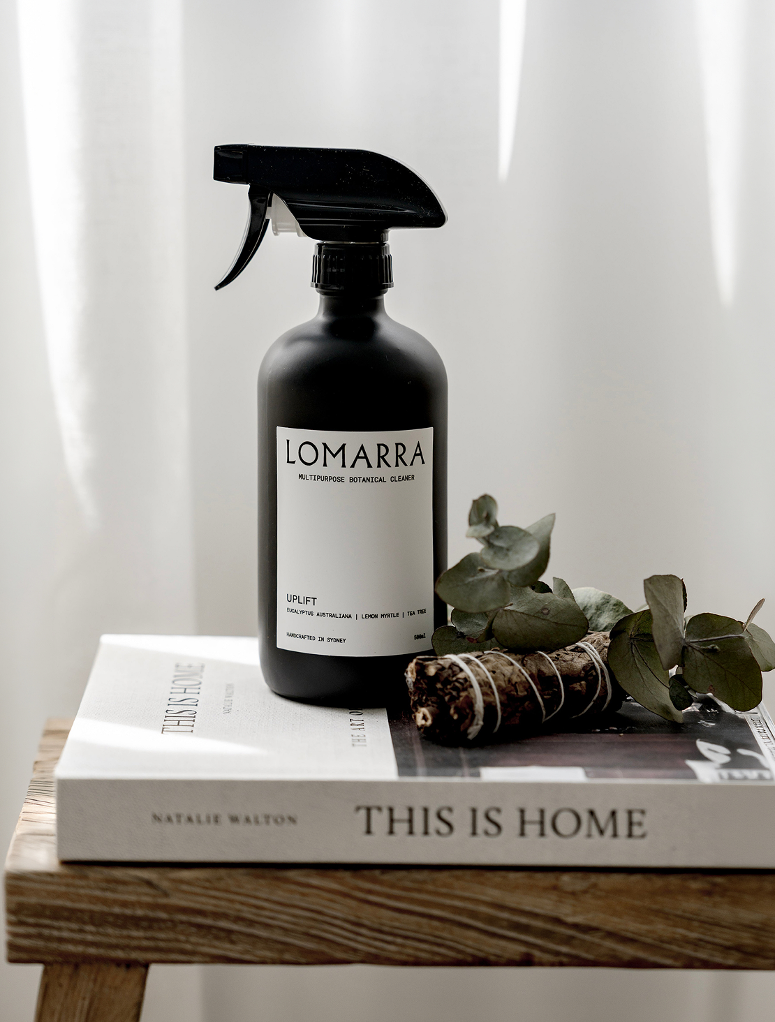 A black spray bottle labeled Lomarra multipurpose botanical cleaner placed on top of a white book titled "This Is Home" by Natalie Walton, with a small bundle of eucalyptus leaves and a small piece of driftwood beside it. The scene is set on a wooden surface with a soft, light background.