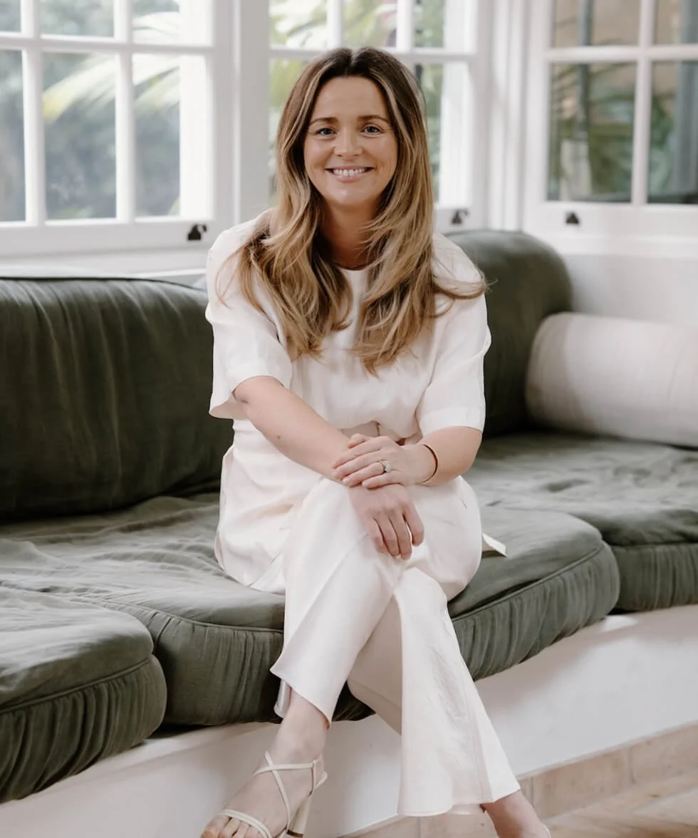 A woman with long wavy brown hair, wearing a white outfit and beige heels, sitting on a dark green couch in a bright room, smiling at the camera.