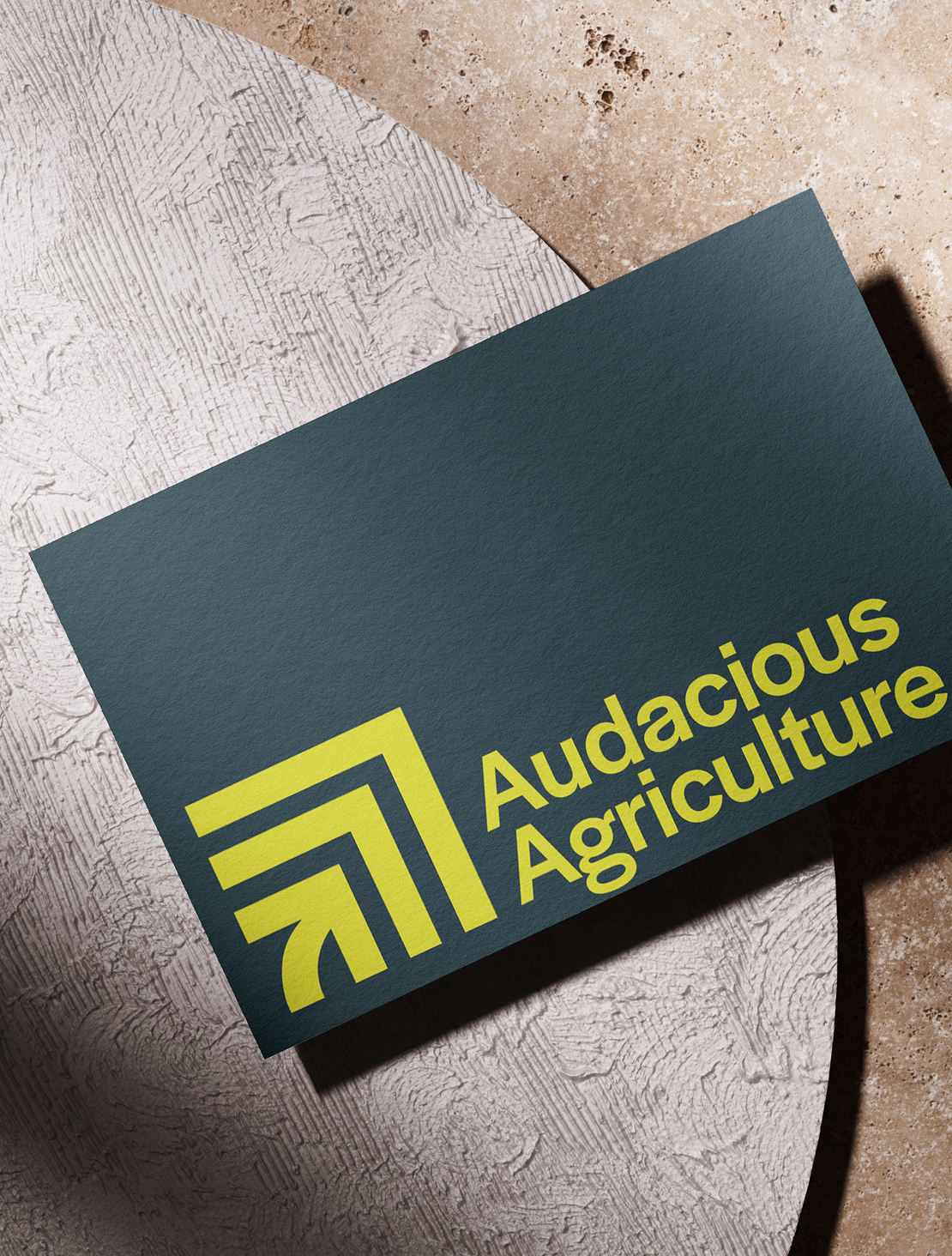 A dark blue business card with yellow text and logo that reads 'Audacious Agriculture' placed on a textured surface with a white circular object underneath.