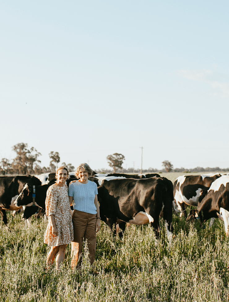 Two women standing in a lush green field with cows grazing around them during daytime.