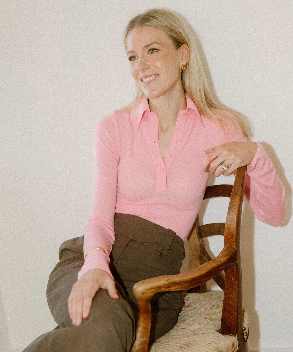 A woman with blonde hair wearing a pink shirt and brown pants, sitting on a wooden chair with light floral upholstery, smiling and looking to her left.