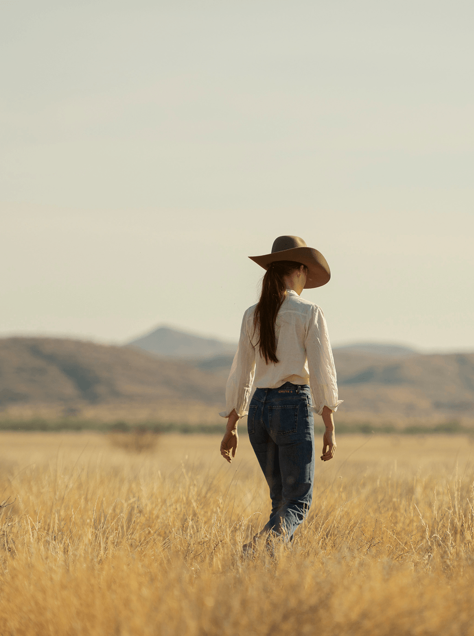 Woman walking through a field of tall grass in a rural landscape with distant hills, wearing a white blouse, jeans, and a wide-brimmed hat.