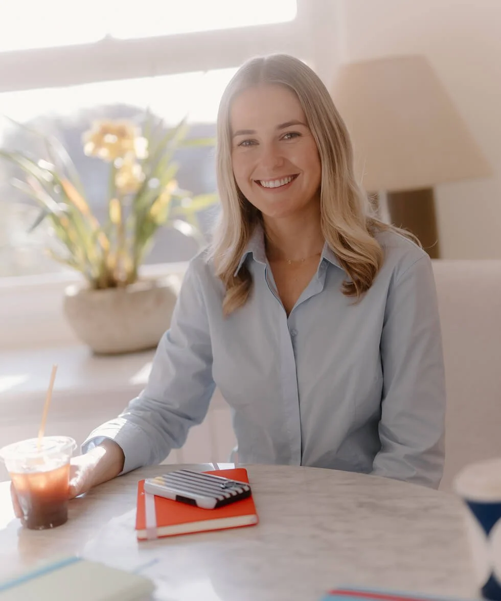 A woman with blonde hair smiling and sitting at a table, with a potted plant and a lamp in the background.