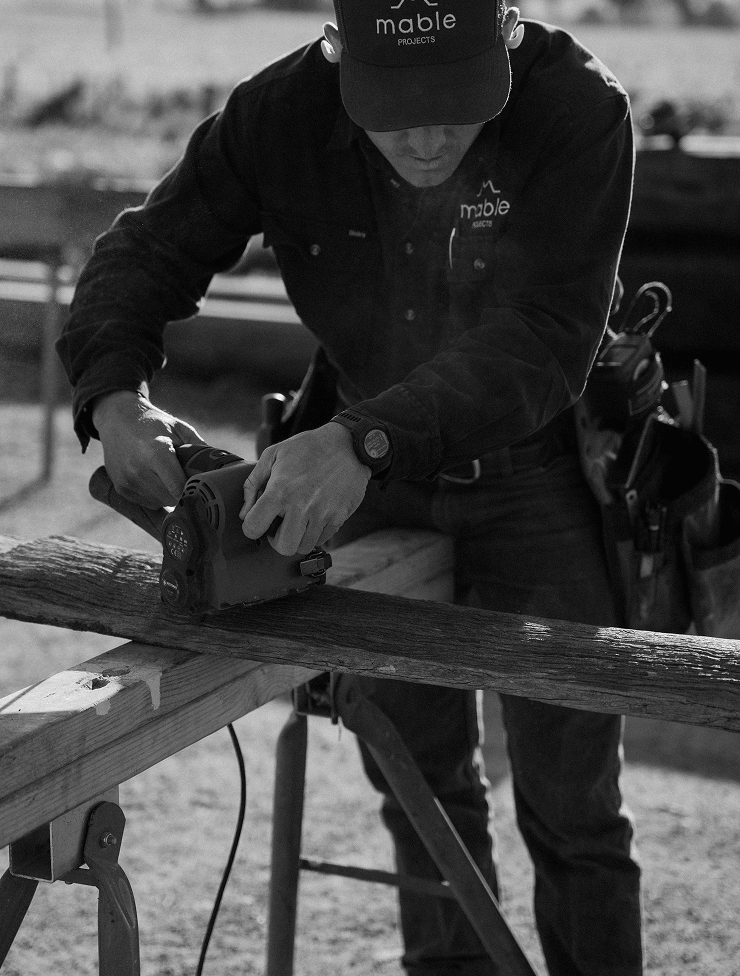 A person wearing a hat and work shirt sawing a piece of wood with a power saw outdoors.