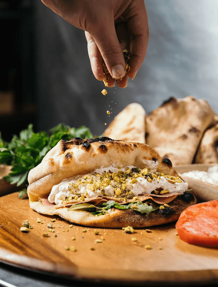 Hand sprinkling crushed pistachios onto a sandwich with bread, lettuce, deli meat, and a spread, on a wooden cutting board.