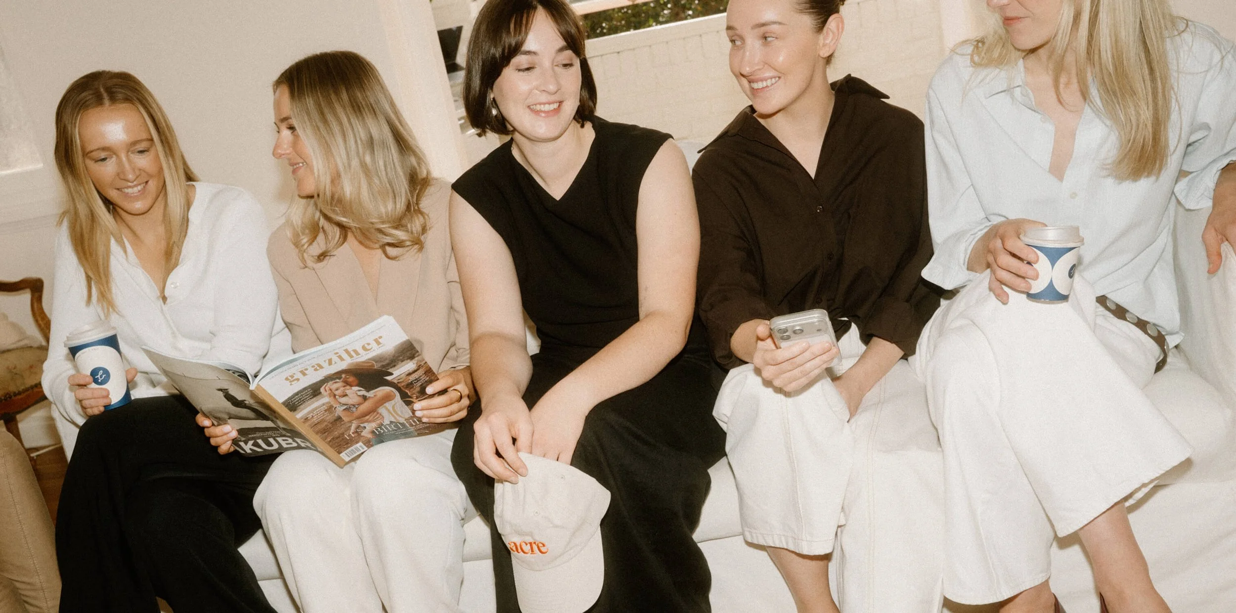Group of five women sitting on a white couch, smiling and chatting, with some holding coffee cups, a magazine, and a phone in a cozy indoor setting.