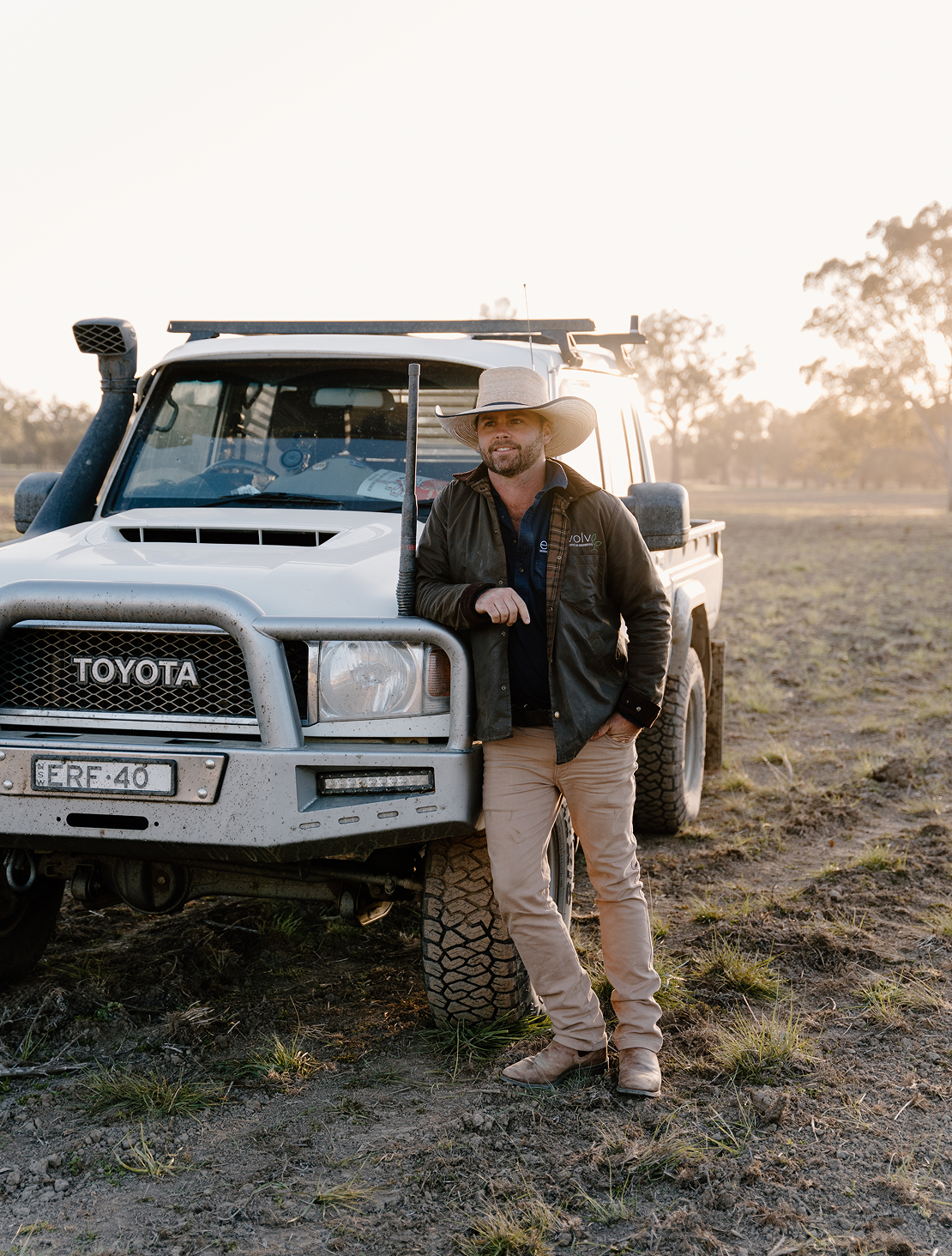 A man standing next to a white Toyota Land Cruiser on a dirt field during sunset, wearing a wide-brimmed hat, dark jacket, and khaki pants.