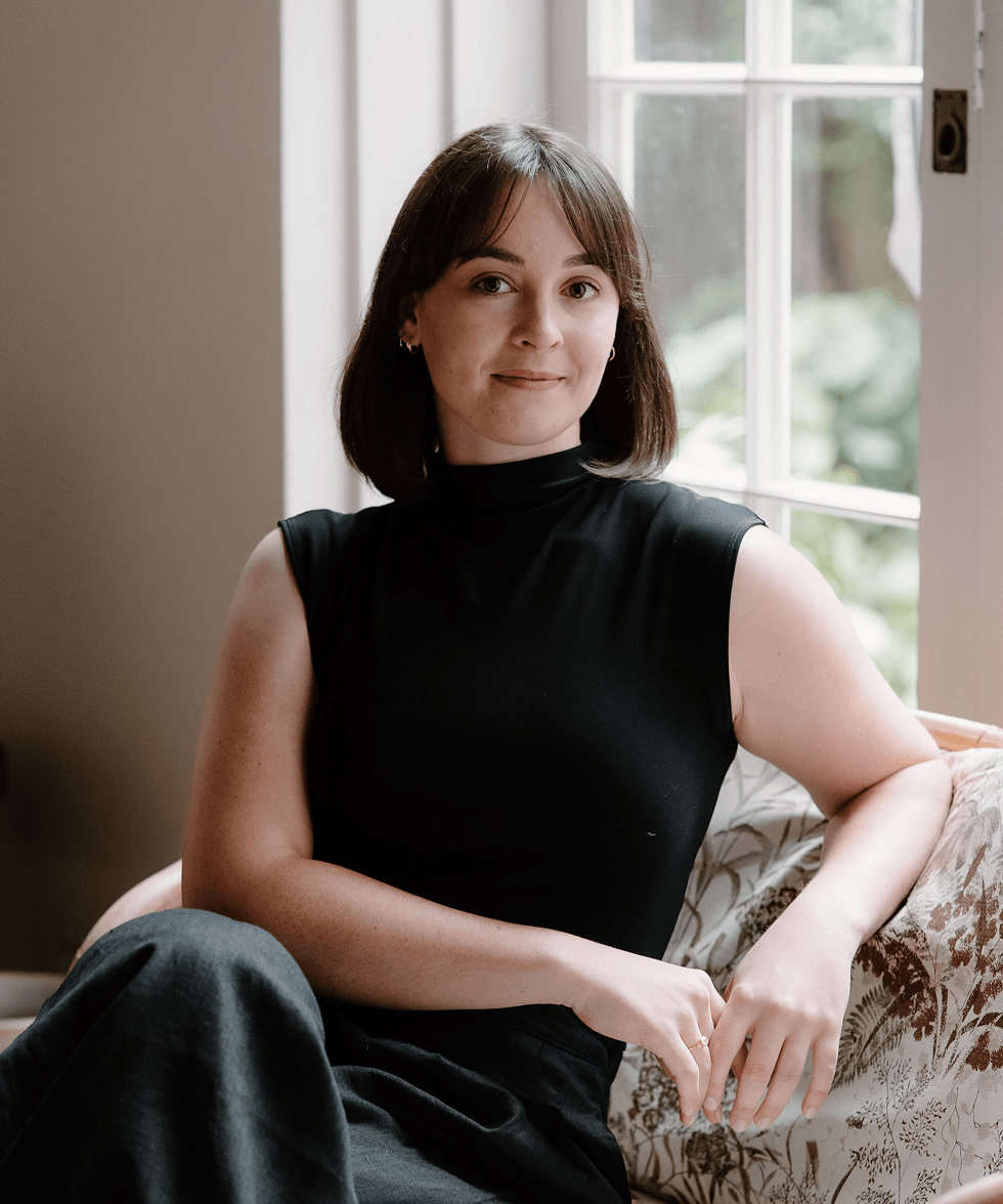 A young woman with shoulder-length brown hair, wearing a sleeveless black top, sitting on a floral-patterned couch near a window in a well-lit room.