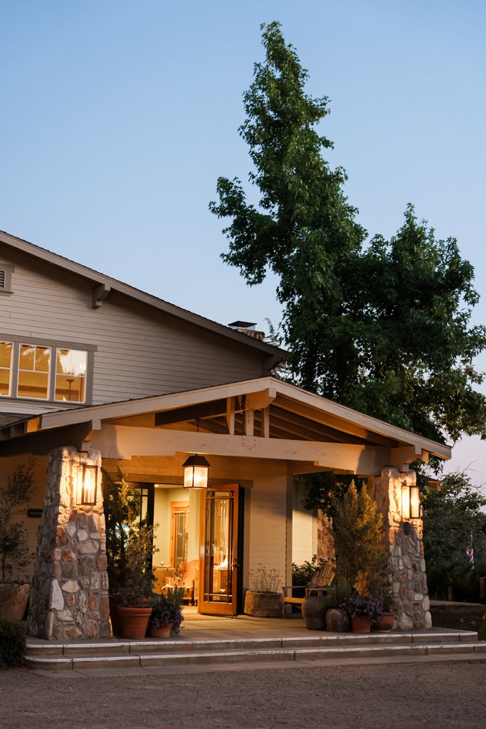 A cozy entrance to Orchard Hill Hotel in Julian CA with stone pillars, potted plants, warm lighting, and a covered porch at sunset.