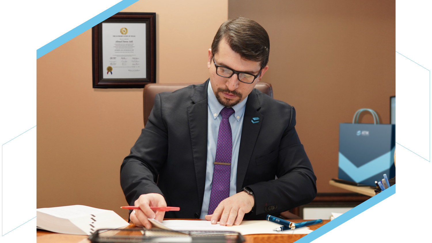 Man in a suit writing at a desk, with framed certificate behind him, and ATN-branded items on the table.