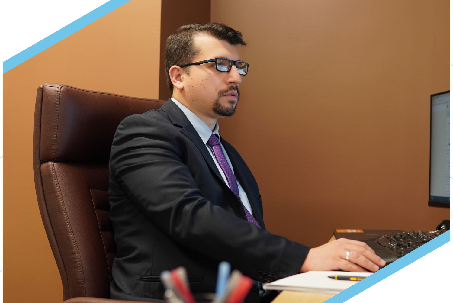 Man in a suit at an office desk using a computer, with glasses and a focused expression.