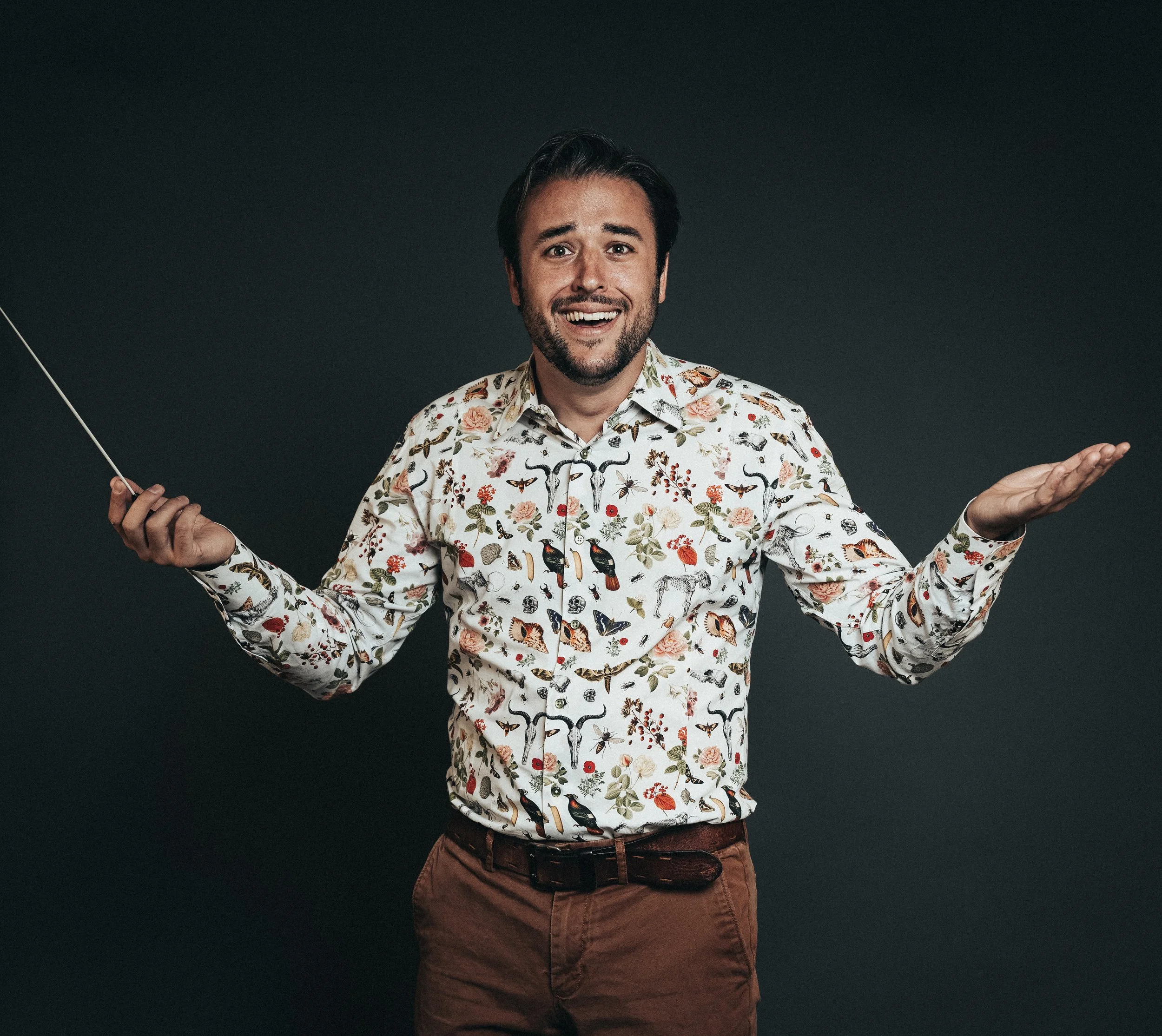 A man with dark hair and a beard wearing a floral patterned shirt and brown pants against a dark background, holding a long thin stick in one hand with a cheerful expression.