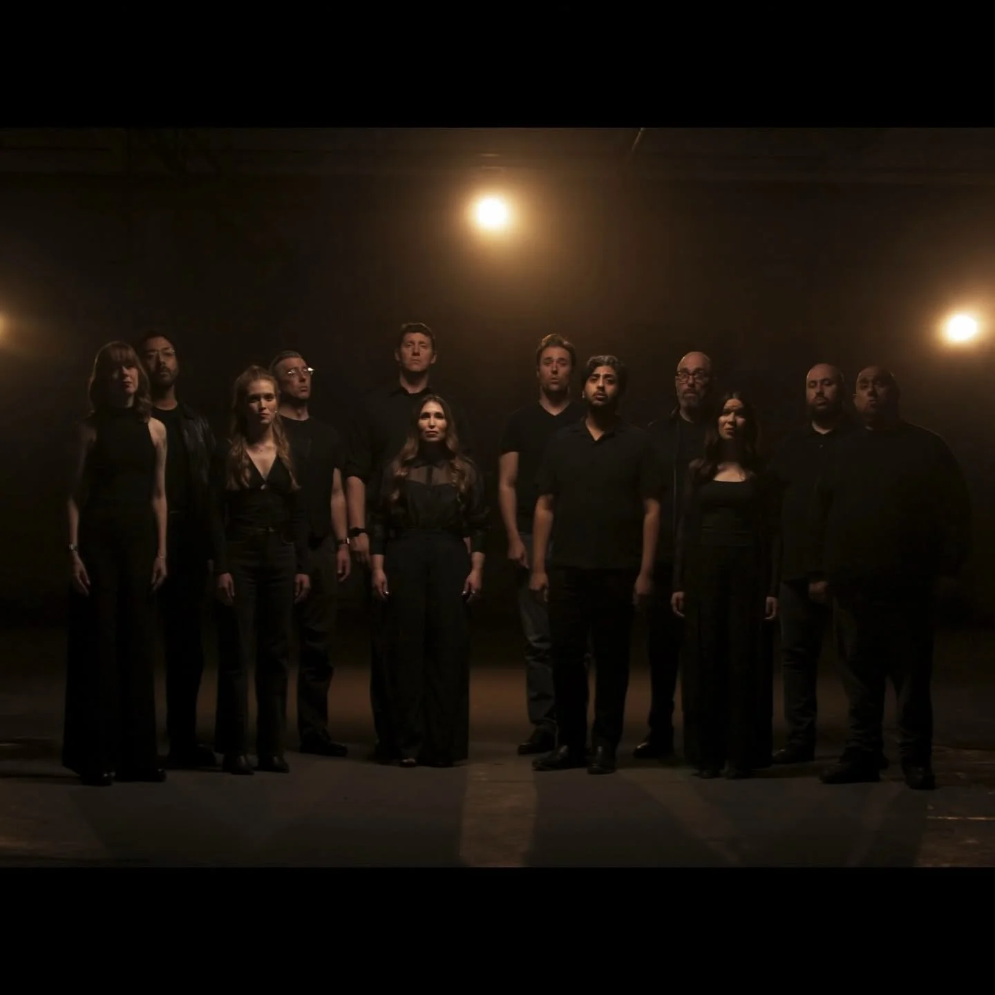 Group of twelve people standing in a dark room under warm lighting, all dressed in black, with serious expressions.