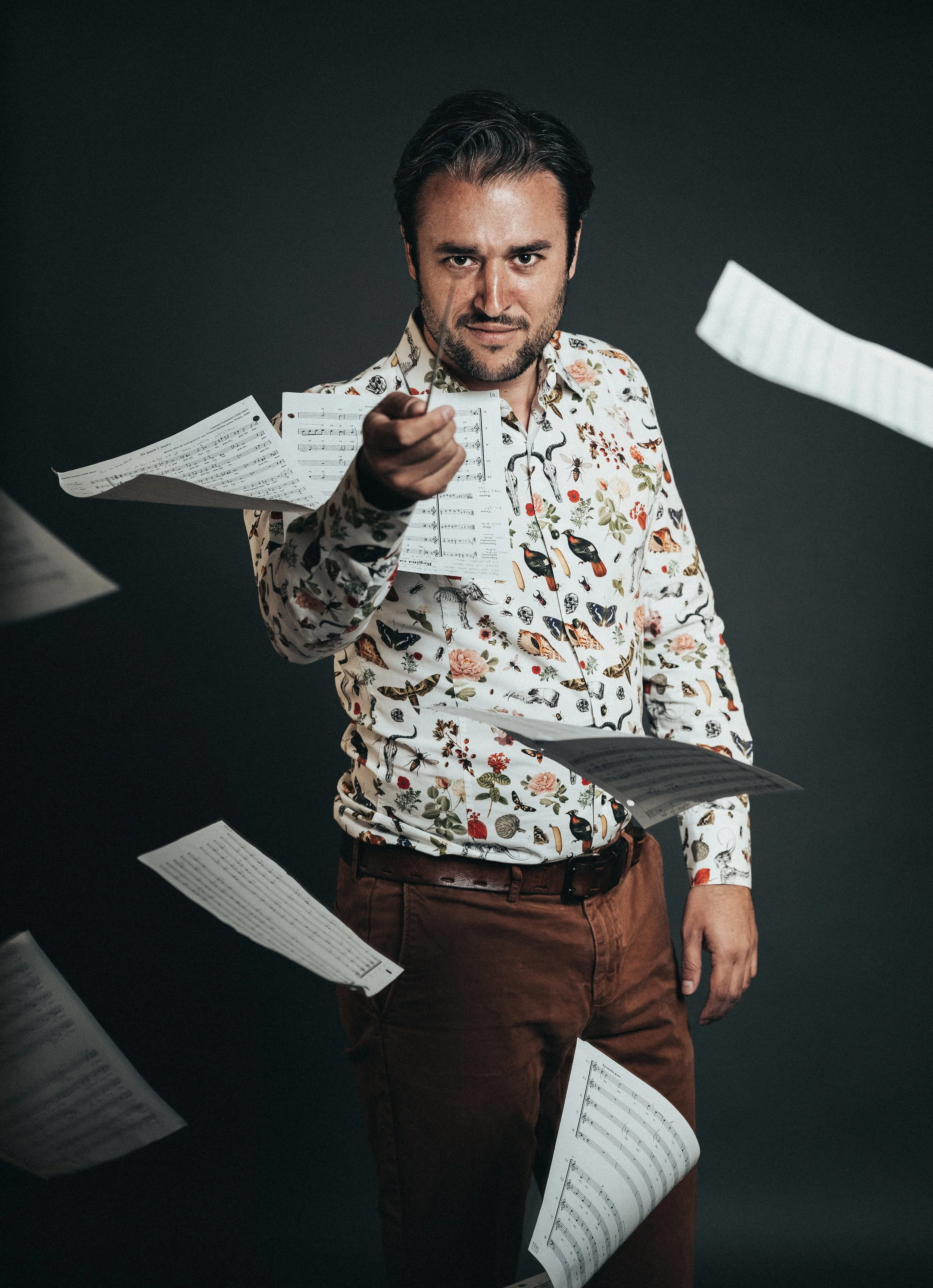 A man with dark hair and a beard in a floral shirt pointing directly at the camera, with scattered sheets of music floating around him against a dark background.