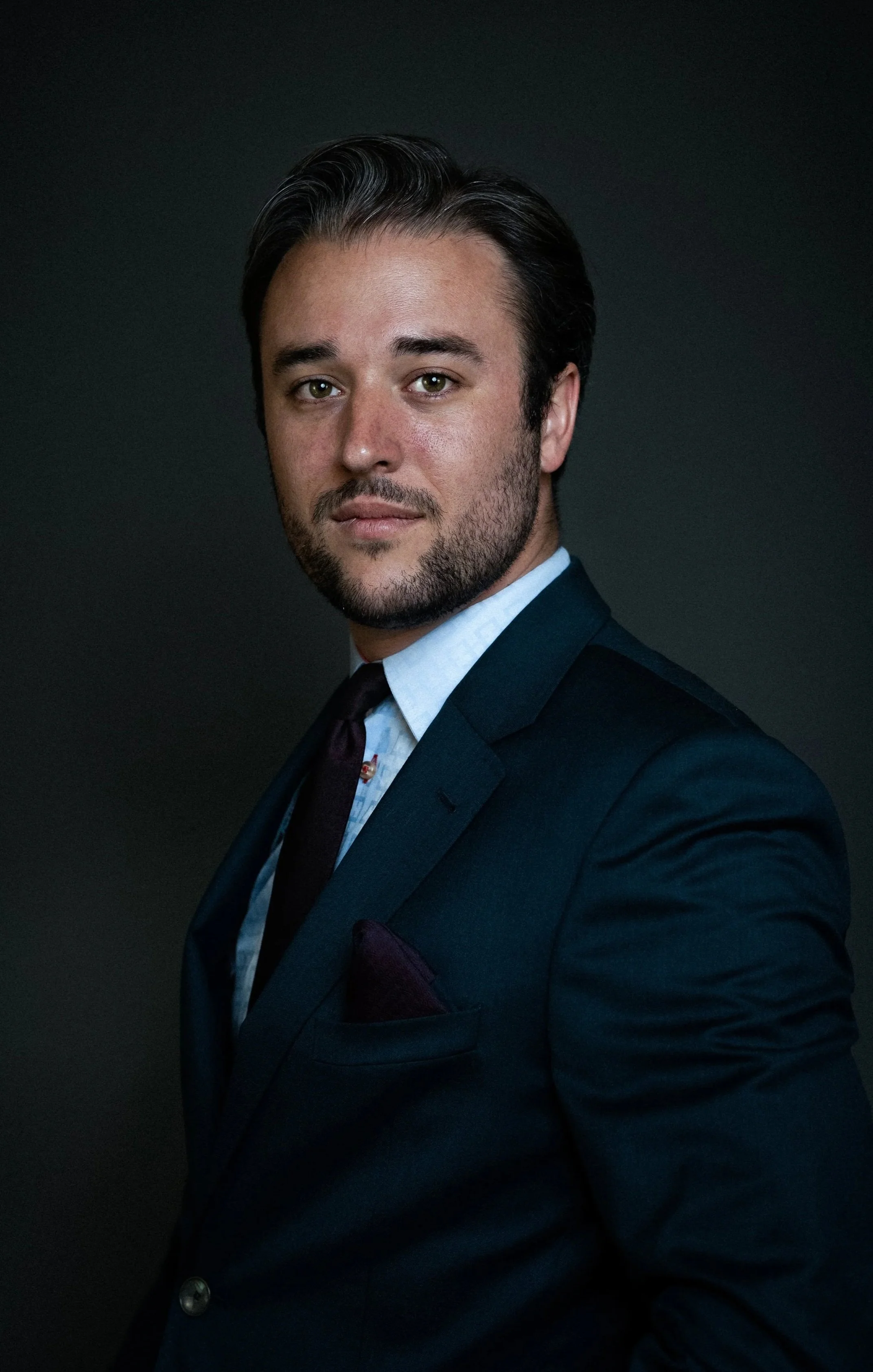 Portrait of a man with dark hair, beard, blue suit, white shirt, dark tie, and pocket square, against a dark background.