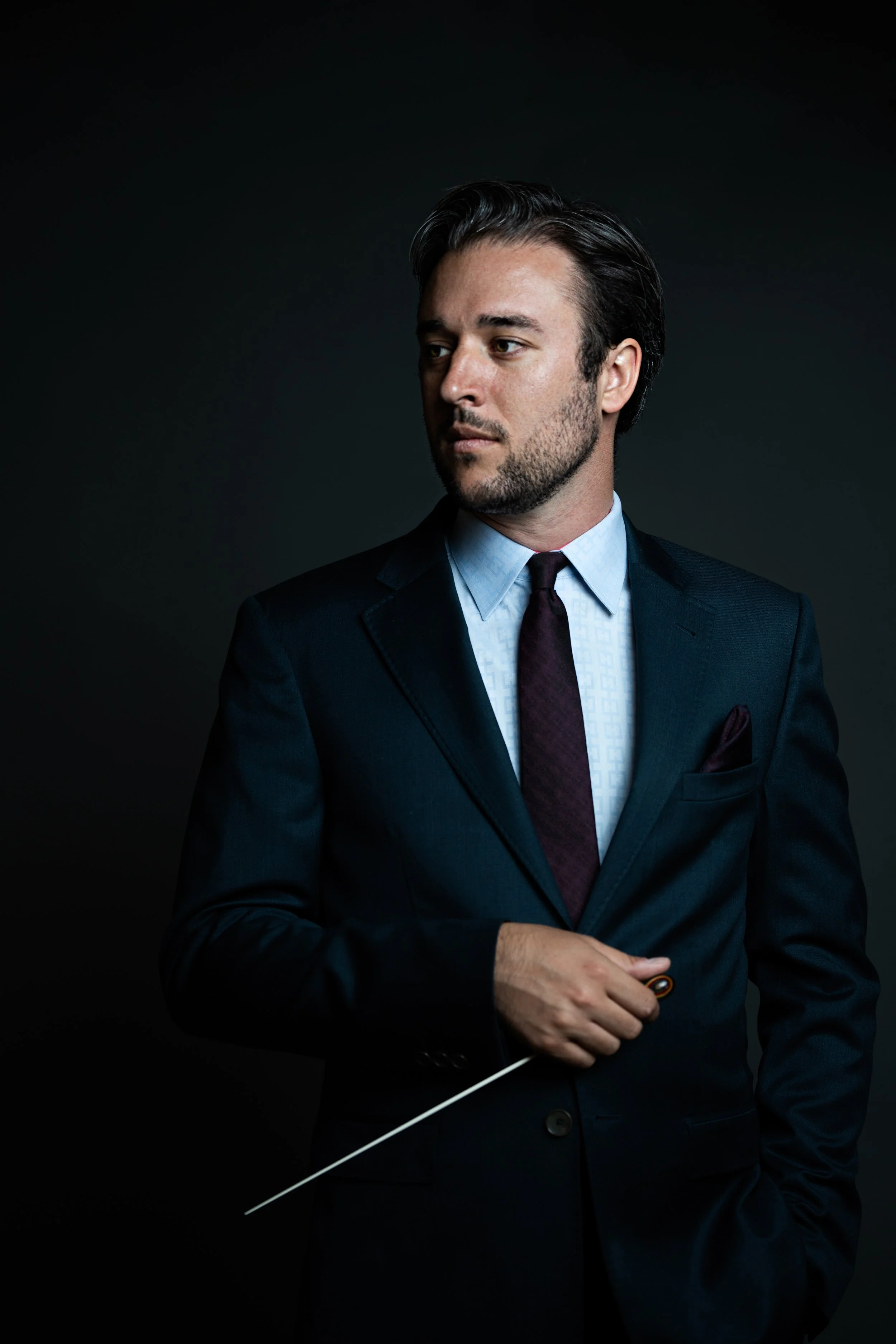 A man with dark hair, dressed in a navy blue suit, white shirt, and maroon tie, holding a pointer stick, and standing against a dark background.