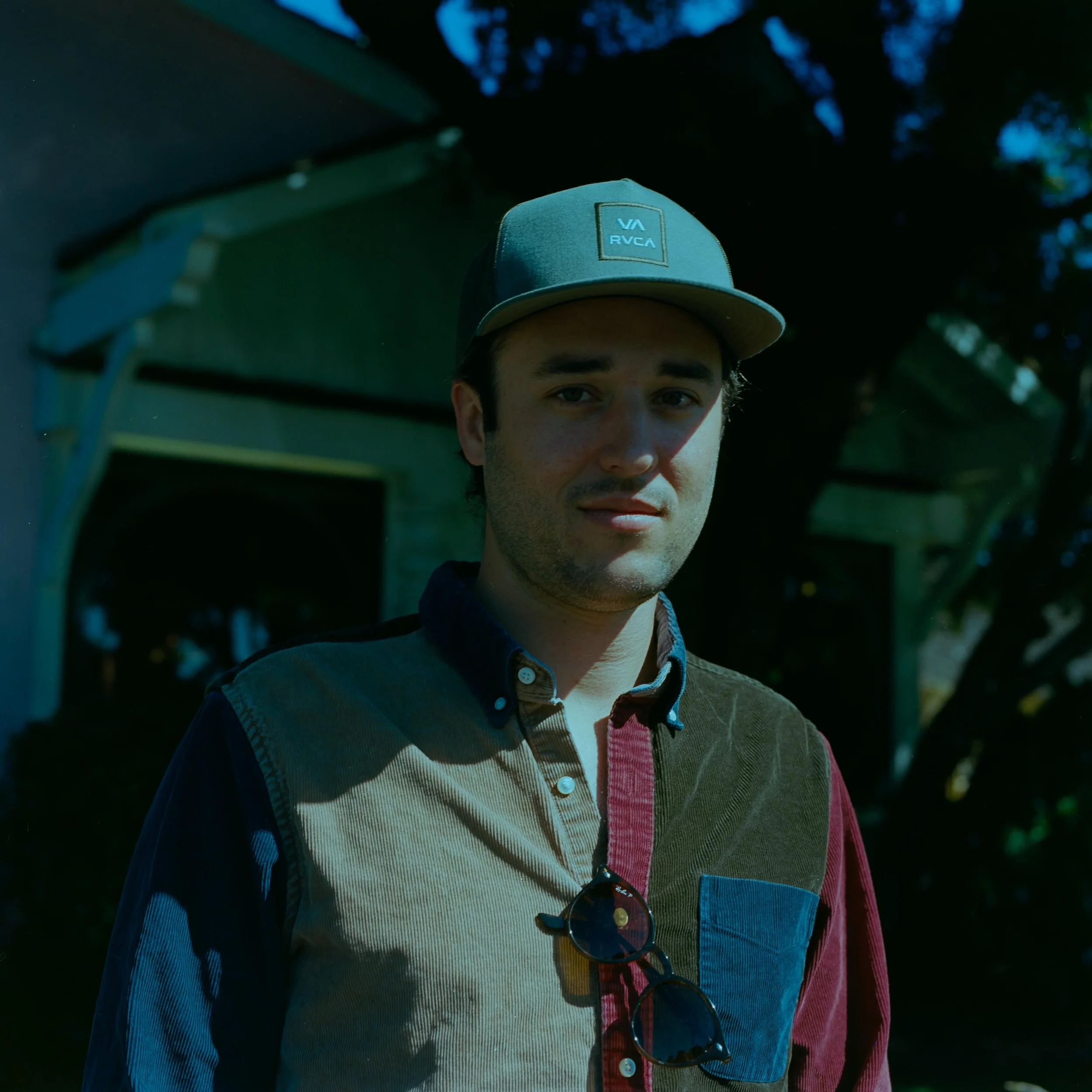 A young man wearing a light blue RVCA baseball cap and sunglasses hanging from his shirt, standing outdoors with trees and a building in the background.